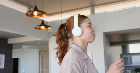 Young woman wearing white headphones enjoying music in modern minimalist apartment kitchen
