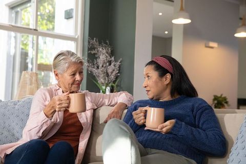 Senior friends chatting on sofa while sipping matching mugs in cozy modern living room