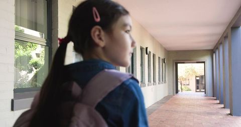 Teenage student standing in campus corridor wearing denim jacket carrying pink backpack backview
