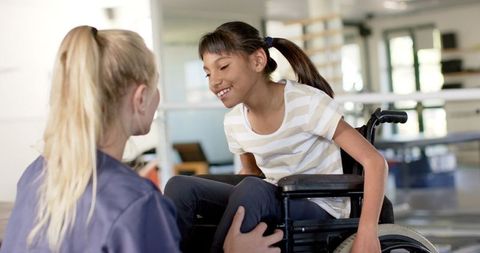 A therapist is assisting a smiling young girl in a wheelchair at a rehabilitation center. The setting highlights compassionate care and support during physiotherapy sessions. This image is ideal for illustrating medical support environments, pediatric health care, inspirational stories about overcoming challenges, healthcare marketing materials, or educational content about rehabilitation services.