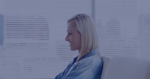 Sitting blonde woman wearing denim jacket facing left in modern lounge with blinds
