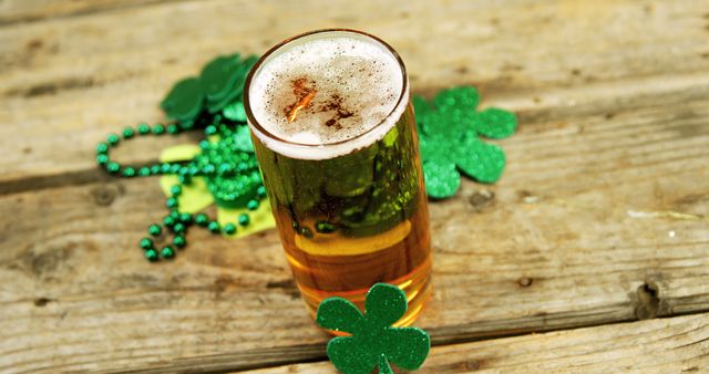 A glass of beer is accompanied by green shamrock decorations and beads, suggesting a celebration of St. Patrick's Day. The rustic wooden surface adds a warm, festive backdrop for the holiday-themed refreshment.