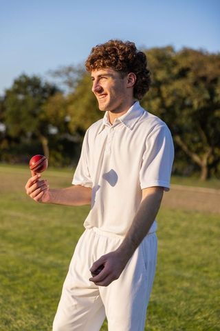 Young cricketer smiling and holding red ball in white uniform on sunlit grassy field