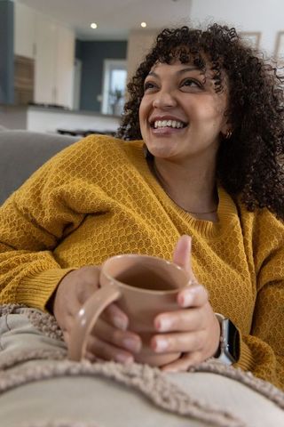 Smiling african american woman reclining on sofa wearing mustard sweater holding warm mug