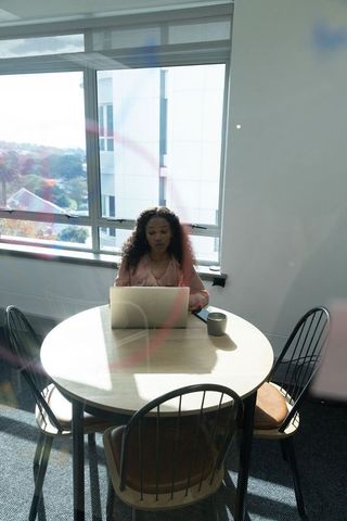 African american woman working on laptop at sunlit round table in modern office