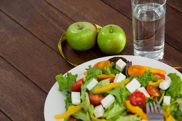 This image depicts a healthy diet concept with a fresh salad, two green apples, a measuring tape, and a glass of water on a wooden table. The salad includes lettuce, cherry tomatoes, yellow bell peppers, and feta cheese. It is ideal for use in articles, blog posts, or promotional materials focused on nutrition, weight loss, healthy living, and fitness.