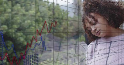 Woman leaning against reflective glass overlaying rising financial charts, appearing pensive