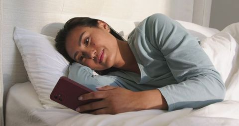 Asian woman lying in bed holding smartphone and looking relaxed in cozy bedroom