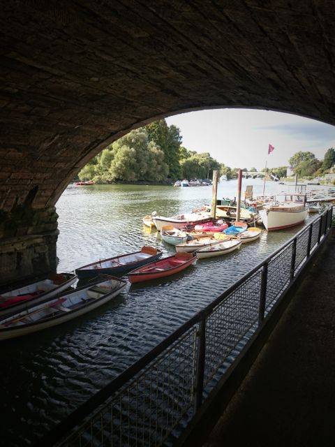 View from underneath a bridge of a scenic riverside dock lined with various rowboats and small boats moored along the side. Calm water reflects the boats, with greenery and trees in the background. This image is ideal for use in travel blogs, boating and leisure-related advertisements, illustrative articles, and outdoor adventure promotions.