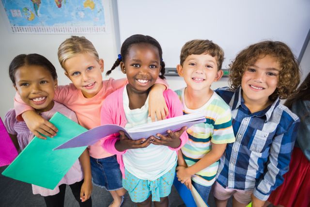This image shows a diverse group of happy children standing together in a classroom, holding a book. They are smiling and appear to be enjoying their time at school. This image can be used for educational materials, school brochures, websites, and advertisements promoting diversity and inclusion in education.