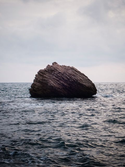 Large rock formation sitting in the middle of a calm ocean, evoking feelings of solitude and peacefulness. Ideal for use in nature-themed designs, inspirational backgrounds, and travel promotions.