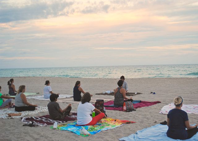A group of people are practicing yoga on a sandy beach during sunset. They are seated on colorful mats, facing the ocean. This can be used to portray themes of wellness, relaxation, outdoor fitness, and tranquility. It is perfect for promoting yoga classes, wellness retreats, beach activities, and peaceful outdoor experiences.