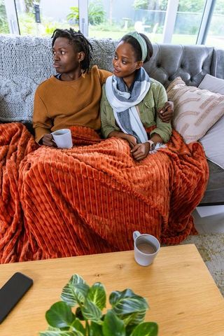 African american couple cuddling on sofa under orange throw, sipping coffee, cozy living room