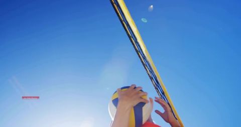 Female players reaching for yellow-blue volleyball at net under bright sun flare