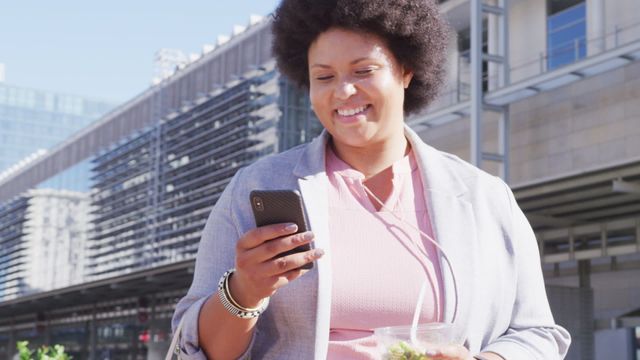 Woman standing on busy street, smiling while using her smartphone. Ideal for use in topics related to digital communication, body positivity, modern lifestyle, urban living, and professional settings.