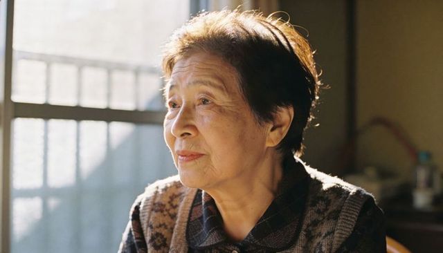 Sunlit senior asian woman gazing out frosted window, contemplative home portrait in soft light