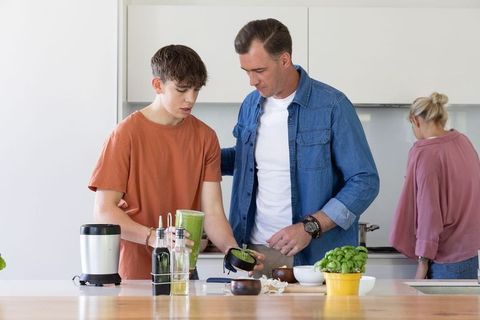 Father guiding teenage son preparing green smoothie at modern kitchen island