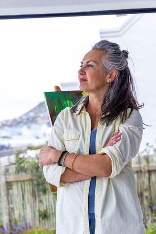 Midlife female artist standing on coastal porch gazing thoughtfully with easel