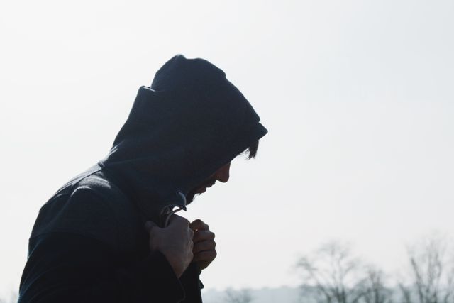 Silhouette of an individual wearing hoodie standing outdoors on a foggy day, possibly feeling deep in thought. Perfect for articles or content involving themes of introspection, solitude, moodiness, or autumn weather. Suitable for mental health awareness campaigns, blogs about solitary activities, or poetic imagery in artistic projects.