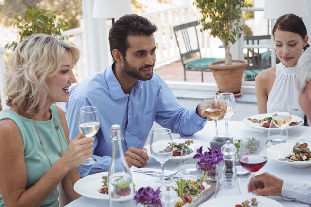 Group of friends enjoying a meal together at an outdoor restaurant. They are engaged in conversation and holding glasses of wine. Ideal for use in content related to social gatherings, dining experiences, friendship, and lifestyle.