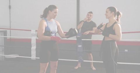 Women exchanging boxing gloves while wrapping hand wraps in ring with coach overseeing training