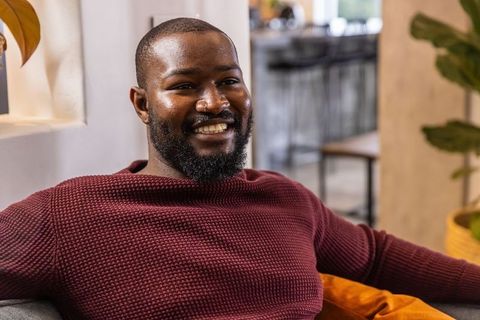 African american man smiling on sofa in cafe wearing burgundy knit sweater, relaxed casual