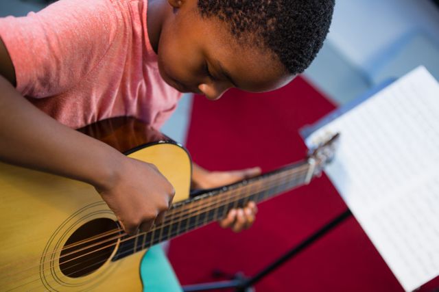 Boy concentrating on playing acoustic guitar in music class. Ideal for educational content, music school brochures, and learning resources for young musicians.