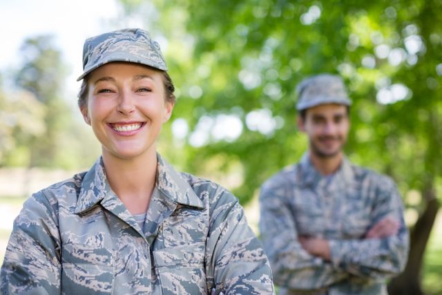 Military couple standing outdoors in park, both wearing camouflage uniforms. Woman in foreground smiling with arms crossed, man in background with arms crossed. Ideal for themes related to military life, teamwork, service, and outdoor activities.
