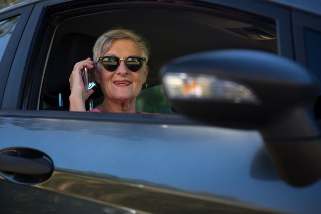 Senior woman wearing sunglasses talking on mobile phone while sitting in car. Ideal for themes related to active aging, communication technology, independence, and modern lifestyle for elderly people.