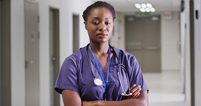 African American female doctor standing in hospital corridor wearing medical attire with a stethoscope around her neck. She appears confident and serious, embodying professionalism and dedication. Ideal for healthcare advertisements, medical articles, patient brochures, online medical content, and educational materials. This visual exudes a strong sense of trust and competence in the healthcare industry.
