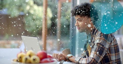 Young man working from home on laptop with winter snow falling outside window
