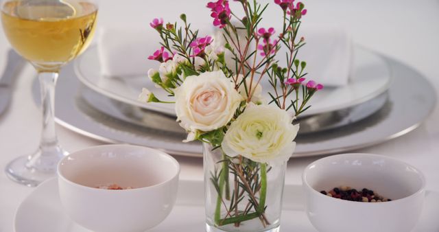 Elegant white table setting featuring a small floral arrangement with pink and white flowers in a clear vase, along with neatly set tableware and a glass of white wine. Ideal for adverts, event planning, restaurant promotions, wedding receptions, romantic dinners, and gourmet dining experiences.