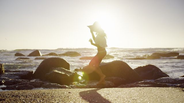 Silhouette of woman walking barefoot on a rocky beach during sunset. Ideal for promoting travel, outdoor adventures, summer holidays, and beach vacations. Can be used for website banners, nature blogs, and relaxation-themed content.