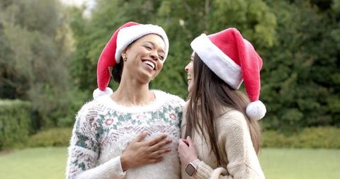 Laughing diverse friends wearing santa hats and cozy knit sweaters sharing holiday cheer outdoors