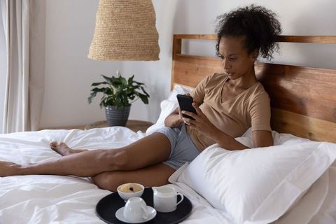 African american woman relaxing on bed using smartphone with breakfast tray, morning light