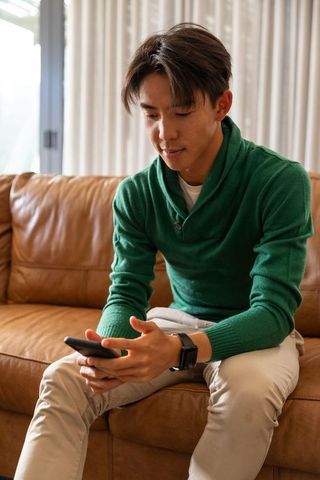 Asian man sitting on leather sofa checking smartphone wearing green sweater and smartwatch