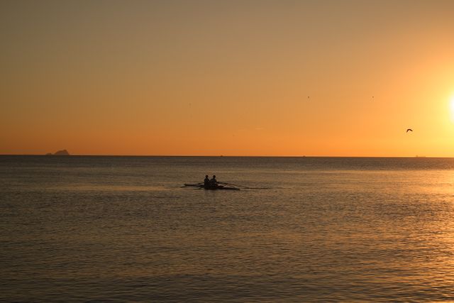 Silhouetted people rowing a boat on a calm ocean during a beautiful sunset. Perfect for themes around tranquility, marine life, outdoor activities, and serene landscapes. Ideal for use in travel promotions, adventure sports materials, and nature-focused content.