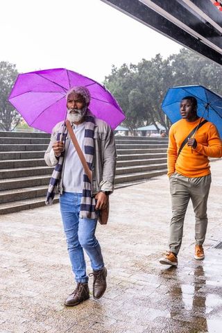 African american men walking in rain with purple and blue umbrellas in urban plaza