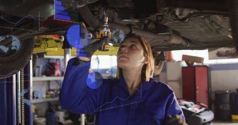 Female mechanic checking undercarriage with cordless drill on car lift in busy garage