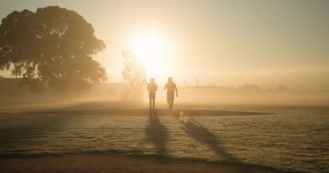 Two friends walking across dewy park at sunrise carrying slender bat, misty silhouettes