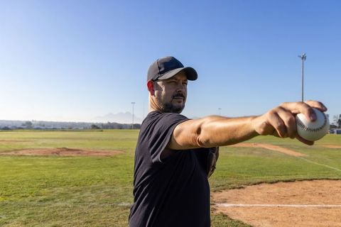 Mature pitcher extending arm holding baseball on sunny ballfield, confident coach portrait