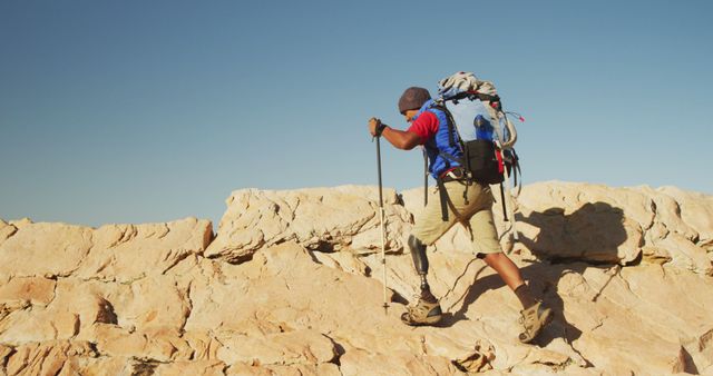 Biracial man with prosthetic leg trekking with backpack and walking poles on rocky mountain. Long distance walking, fitness, challenge, disability, nature and healthy outdoor lifestyle.