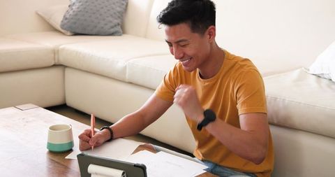 Asian Man Fist Pumping in Living Room with Tablet at Home