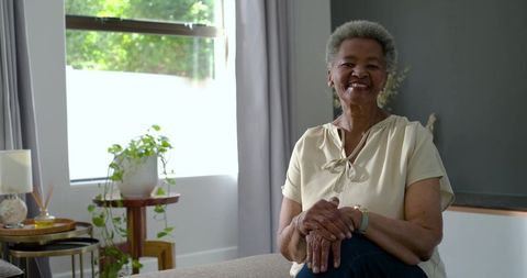 Smiling senior woman sitting on sofa by window in minimalist cozy room, daylight portrait