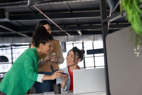 Multicultural female team collaborating in modern office smiling and pointing at smartphone