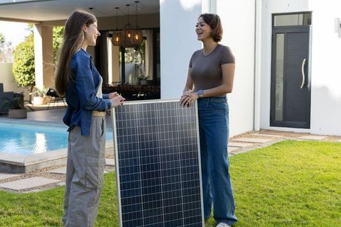 Women holding solar panel and chatting in modern backyard by pool