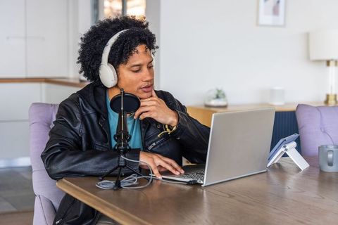 African american podcaster recording at home with laptop, headphones and microphone