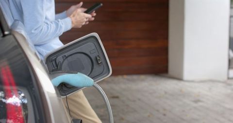 Man charging electric car at home, using smartphone