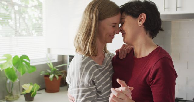 Lesbian couple dancing happily, showing affection and joy in their kitchen. The background has houseplants and kitchen decor, suggesting a cozy home environment. Ideal for portraying loving relationships, LGBT representation, and domestic bliss. Suitable for use in relationship blogs, LGBT advocacy materials, and family-oriented content.