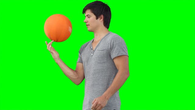 A young man concentrating on spinning a basketball on his finger, set against a green background, emphasizing focus and control. Useful for sports training, talent showcases, or motivational content. Perfect for advertisements promoting athletic gear, basketball training techniques, or competitions due to its dynamic composition and bright colors.